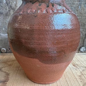 Puede incluir: Una gran olla de terracota hecha a mano con un cuerpo ancho y redondeado y un borde ligeramente ensanchado. La olla presenta una combinación de colores de dos tonos, con una base marrón rojizo más clara y una sección superior marrón más oscura y brillante. Se ven detalles decorativos tallados cerca del borde.