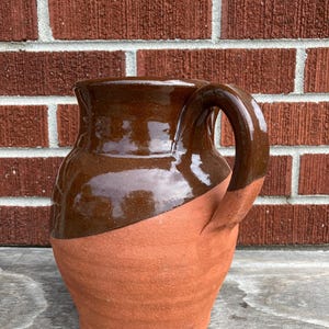 Może przedstawiać: Ceramiczny dzbanek o dwukolorowym wzorze, z gładką, błyszczącą ciemnobrązową glazurą w górnej części oraz teksturowanym, matowym wykończeniem z gliny w kolorze terakoty w dolnej części, wyposażony w zakrzywiony uchwyt po prawej stronie.
