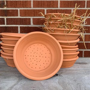 May include: Several terracotta plant saucers and pots are displayed. The saucers have drainage holes in the centre. The pots are stacked, some with dried straw. The background includes a red brick wall and a weathered wooden surface.