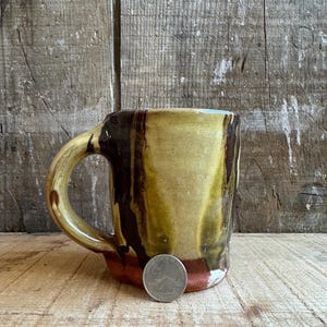 May include: Handmade ceramic mug with a unique design. The mug features a blend of brown, yellow, and red hues. It has a sturdy handle and a quarter coin is next to the mug for size reference. The mug is on a wooden surface.
