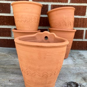 May include: A collection of terracotta flower pots in various sizes and shapes. The pots have a natural, earthy tone and feature a wavy, etched design around the middle. One pot has a flat back with a hole for hanging. The pots are stacked on a wooden surface.