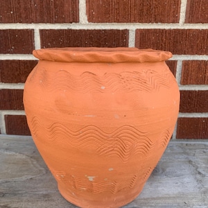 May include: A terracotta-colored, round, ceramic pot with a lid. The pot features a wavy, textured design around the body and a scalloped edge on the lid. The pot is sitting on a wooden surface.