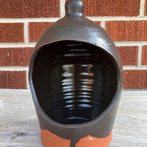 Puede incluir: Un salero de cerámica de color marrón oscuro con un interior brillante y una abertura ancha y arqueada. El exterior presenta una sección superior esmaltada en marrón oscuro liso que se transforma en una base de terracota mate sin esmaltar.