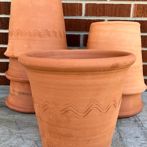 May include: Several terracotta flower pots of varying sizes are arranged against a brick wall. The pots have a classic, slightly flared shape with a decorative wavy line etched around the middle. The unglazed clay has a warm, earthy tone.