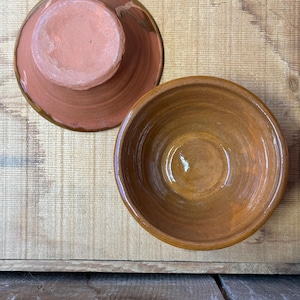 May include: Two handmade terracotta bowls. One bowl is upside down, revealing a pinkish-brown base and a darker rim. The other bowl is right-side up, displaying a warm brown interior. Both bowls sit on a wooden surface, showcasing their rustic charm.