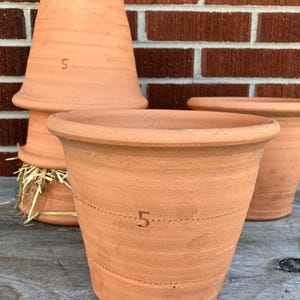 May include: Several terracotta flower pots of varying sizes are displayed. The pots have a classic, slightly flared shape with a rounded rim. Each pot is a warm, earthy orange-brown color and features the number "5" stamped on the side.