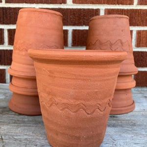Op de afbeelding: Een verzameling terracotta bloempotten. De potten hebben een golvend, geëtst ontwerp in het midden. De potten zijn gestapeld en gerangschikt voor een bakstenen muur.