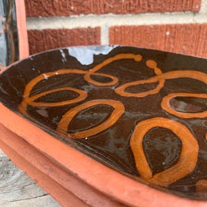 May include: A rectangular ceramic tray with a dark brown glazed interior featuring a light orange swirling line pattern. The exterior of the tray is unglazed terracotta with a visible rim and a smooth, slightly curved edge design.