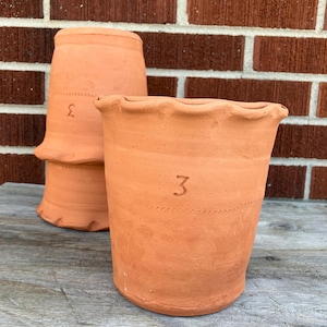 May include: Two terracotta flower pots. One pot has a scalloped rim and the number "3" written on the side. The other pot is upside down behind the first pot and also has the number "3" written on the side. The background is a red brick wall.