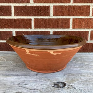 May include: A wide, circular ceramic bowl with a smooth, dark brown glazed interior and rim. The exterior features a natural, unglazed terracotta finish with a light-colored, irregular horizontal band circling the upper portion of the vessel.