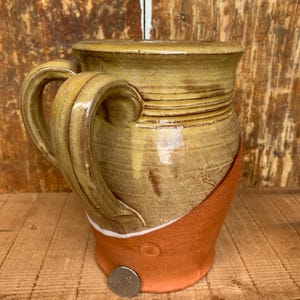 Op de afbeelding: Een handgemaakte aardewerken mok met twee handvatten, met een bruine en gele glazuur. De mok heeft een uniek ontwerp met een contrasterende oranje basis.