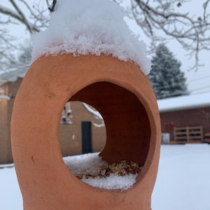 May include: A terracotta bird feeder, covered in snow, hangs from a black wire. The feeder has a large circular opening and is filled with birdseed and snow. The background shows a snowy outdoor scene.