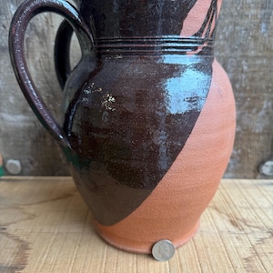Op de afbeelding: Handgemaakte keramische kan met een grote handgreep. De kan heeft een donkerbruin geglazuurde bovenkant en een diagonale terracotta-kleurige sectie. De basis is ongeglazuurd. De kan staat op een houten oppervlak.