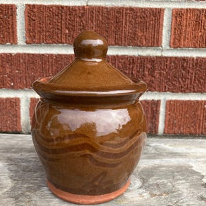 May include: A small, round, brown glazed ceramic jar with a matching lid. The jar features a glossy finish with subtle, darker horizontal wavy bands circling the body. The base of the jar is unglazed, revealing the natural reddish-brown clay color.
