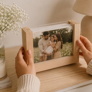 Può includere: Una cornice rettangolare in legno chiaro contenente la fotografia di una famiglia di tre persone in un campo. La cornice presenta un bordo spesso in legno naturale con un pannello in acrilico o vetro trasparente a protezione.