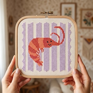Peut inclure: Un tambour à broder carré en bois contenant un motif au point de croix représentant une crevette orange et rouge avec un visage souriant. La crevette est centrée sur un fond à rayures verticales violet clair sur toile blanche.