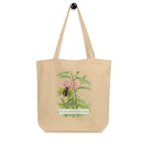 Può includere: Una borsa tote beige con una grafica rettangolare centrata raffigurante un bombo appoggiato su un grappolo di piccoli fiori rosa con foglie verdi. Sotto l'illustrazione, il testo "Bumblebee's Delight" è stampato in un carattere verde scuro.