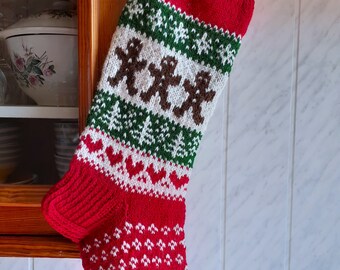 May include: A festive Christmas stocking, knitted in red, white, and green. The design features gingerbread men, trees, and hearts. The stocking is hanging on a wall. The top and toe are red, with a patterned section in the middle.