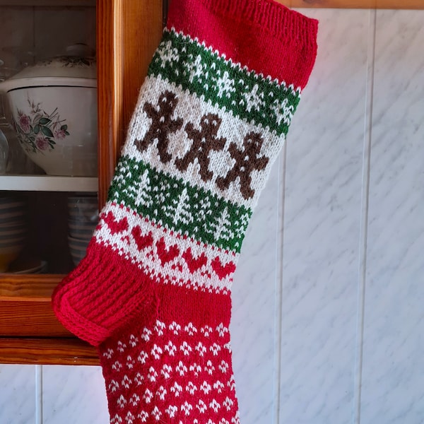 May include: A festive Christmas stocking, knitted in red, white, and green. The design features gingerbread men, trees, and hearts. The stocking is hanging on a wall. The top and toe are red, with a patterned section in the middle.