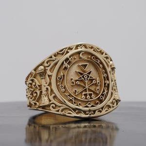 May include: A gold-toned ring featuring an oval face with an engraved occult sigil, including a central star, triangle, and numerical symbols. The band is decorated with intricate scrollwork and a small pillar motif on the side of the ring.