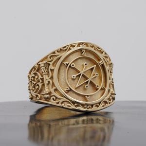 May include: A gold-toned ring featuring a circular face with a central sigil design surrounded by the letters Z, A, E, and A. The band and sides of the ring are decorated with intricate, raised scrollwork and pillar-like motifs in a metallic finish.