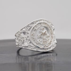 May include: A silver ring featuring an oval face with an intricate engraved sigil and a five-pointed star. The band displays detailed scrollwork and relief carvings of architectural elements, including a tower and a lantern on the sides.