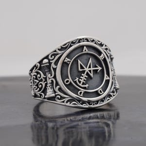 Puede incluir: Un anillo de plata con acabado oxidado y volutas intrincadas en la banda. La cara circular muestra un sello central con las letras A, B, A, D, O, N dispuestas alrededor del perímetro, acentuado por un pequeño motivo de columna.