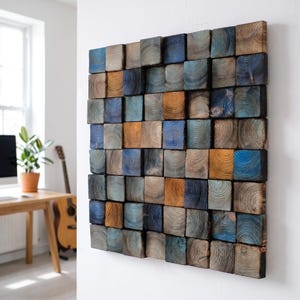 May include: A square wall art piece composed of numerous small, individual wooden blocks arranged in a grid. Each block features visible circular wood grain patterns, with a colour palette consisting of shades of blue, brown, and natural wood tones.