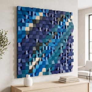 Puede incluir: Una pieza de arte mural cuadrada compuesta por numerosos bloques de madera individuales dispuestos en una cuadrícula. Los bloques están pintados en varios tonos de azul, azul marino oscuro y crema claro, creando una superficie geométrica tridimensional texturizada.