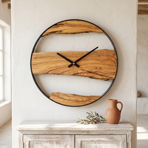 May include: A circular wall clock featuring a black metal frame and two horizontal segments of natural light brown wood with visible grain patterns. The clock has two thin black hands centred on the wood, mounted on a white wall.
