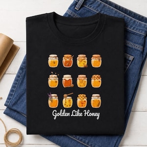 May include: A black crew neck t-shirt featuring a graphic print of twelve glass jars filled with various shades of golden honey, some with wooden dippers, and the white text "Golden Like Honey" printed underneath the jars.