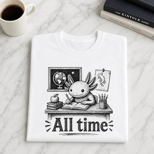 May include: A white t-shirt featuring a black and white graphic of an axolotl sitting at a desk, writing in an open book. The desk includes a globe, an apple, and a pencil holder. The text "All time" is printed in large letters below the desk.