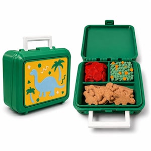 May include: A green plastic lunch box with a white handle and a yellow panel featuring a blue dinosaur and palm tree graphic. The open lunch box reveals three compartments containing red, green, and brown textured food-shaped items.