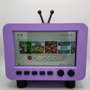 May include: A purple television-shaped stand housing a Nintendo Switch console. The screen displays the home menu with game icons for Luigi's Mansion 3, Animal Crossing, Mario Kart, and Nintendo Switch Sports. It features black knobs, buttons, and a V-shaped antenna.