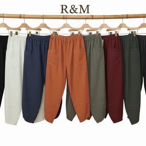 May include: A collection of eight pairs of trousers hanging on wooden hangers from a bamboo rod. The trousers are available in black, white, navy, orange, olive green, burgundy, dark green, and charcoal grey, each featuring an elastic waistband.