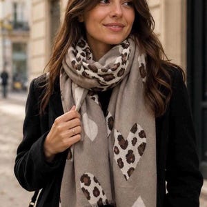 May include: A beige scarf featuring a repeating pattern of large hearts with a brown and black leopard print design. The scarf has a soft texture, a rectangular shape, and frayed edges along the bottom hem, worn wrapped around the neck.