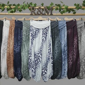 May include: A collection of eleven loose-fitting harem trousers hanging on a wooden rack with the letters R&M displayed at the top. Each pair features a wide, solid-coloured fold-over waistband and a distinct animal print pattern in various colours.