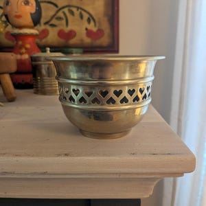 Puede incluir: Un cuenco de latón pulido con un borde ancho y acampanado, y una banda decorativa de corazones recortados que rodea la parte central. El metal tiene un tono dorado cálido con un acabado liso y reflectante, y una base redondeada.