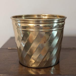 May include: A polished brass planter featuring a cylindrical shape with a distinct diagonal ribbed or fluted pattern along the exterior surface. The metal has a reflective gold-toned finish with a smooth, rounded rim at the top edge.