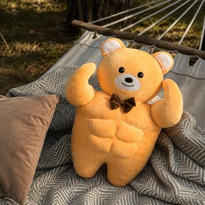 May include: A plush toy shaped like a muscular bear with golden yellow fur, a white muzzle, and black eyes. The bear features a defined six-pack abdominal area, flexed arms, and a small brown bow tie attached to its neck. The toy has rounded ears.