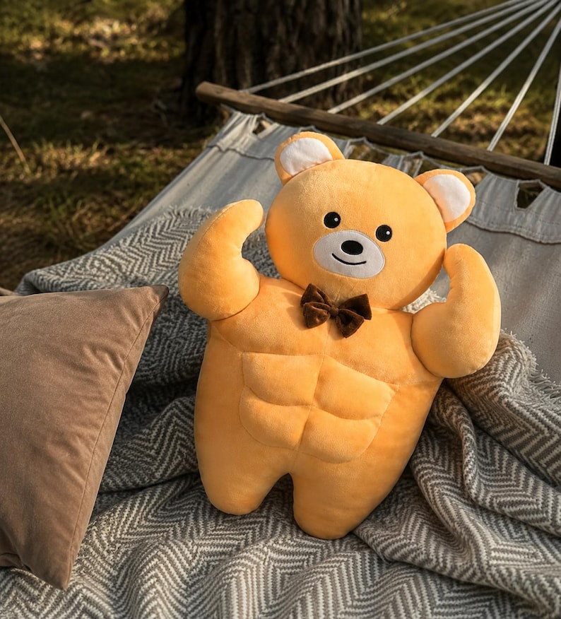 May include: A plush toy shaped like a muscular bear with golden yellow fur, a white muzzle, and black eyes. The bear features a defined six-pack abdominal area, flexed arms, and a small brown bow tie attached to its neck. The toy has rounded ears.