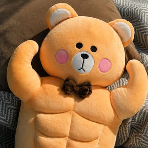 May include: A plush toy shaped like a bear with orange fur, featuring a muscular torso with defined abdominal muscles, pink circular cheeks, a white muzzle, a black nose and mouth, and a small brown bow tie attached at the neck area.