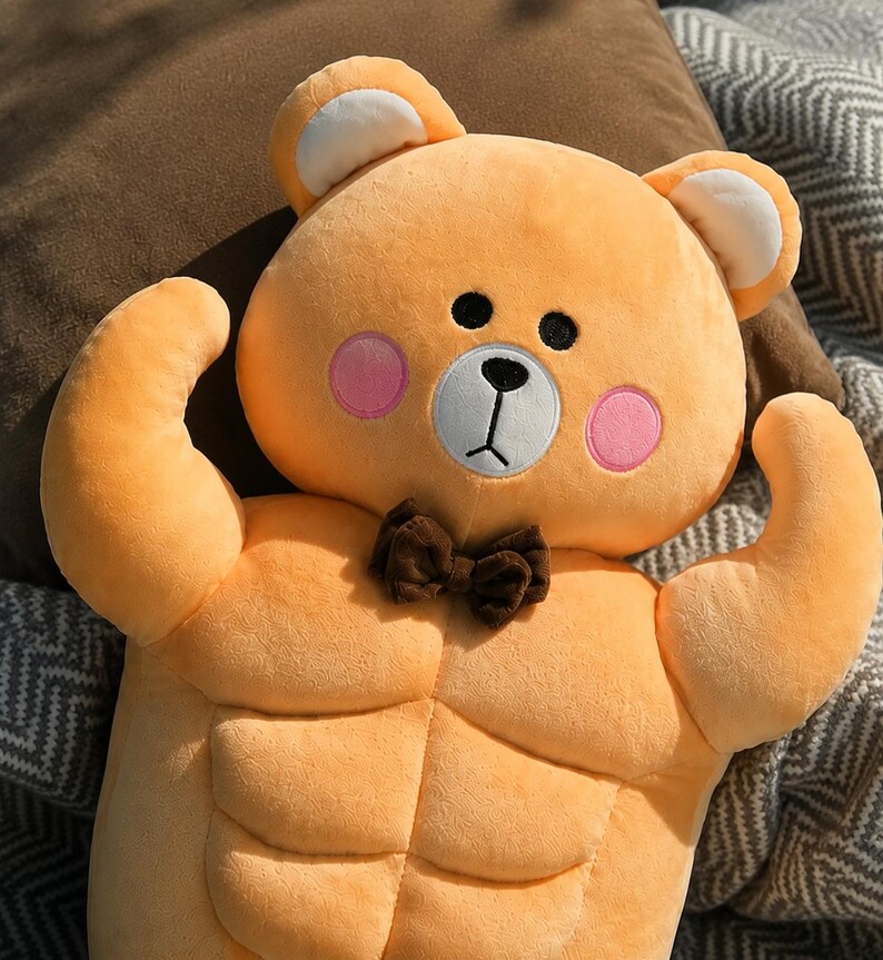 May include: A plush toy shaped like a bear with orange fur, featuring a muscular torso with defined abdominal muscles, pink circular cheeks, a white muzzle, a black nose and mouth, and a small brown bow tie attached at the neck area.