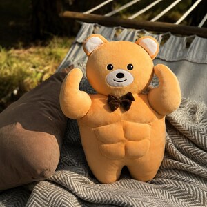 May include: A plush toy shaped like a light orange bear with a muscular torso, defined abdominal muscles, and raised arms. The bear features a white muzzle, black eyes, a small black nose, and a dark brown bow tie attached at the center of the chest.