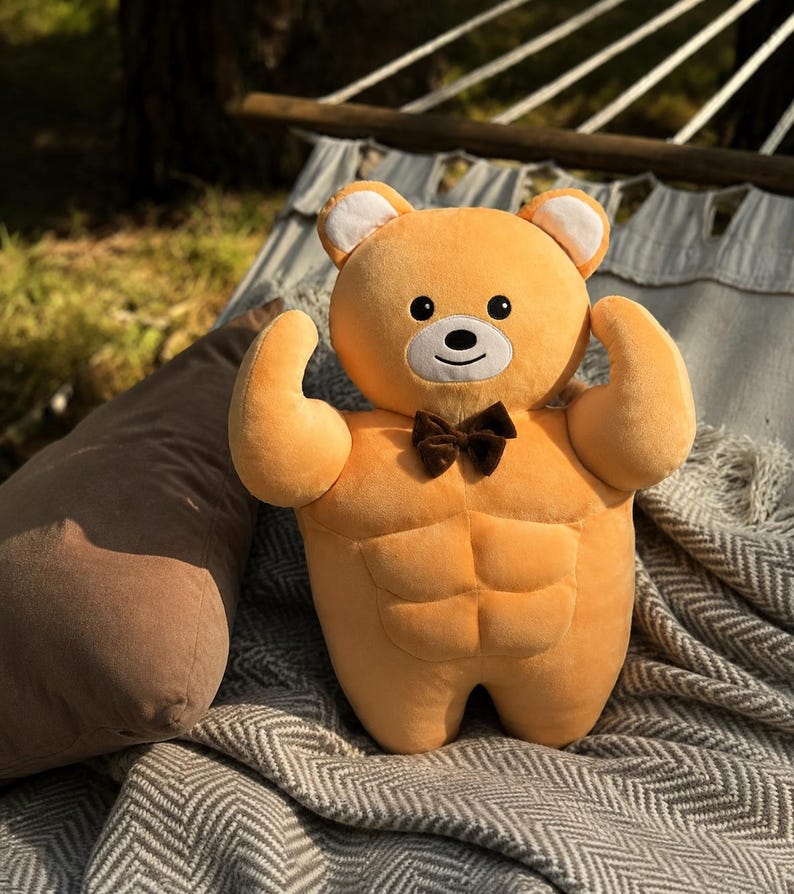 May include: A plush toy shaped like a light orange bear with a muscular torso, defined abdominal muscles, and raised arms. The bear features a white muzzle, black eyes, a small black nose, and a dark brown bow tie attached at the center of the chest.