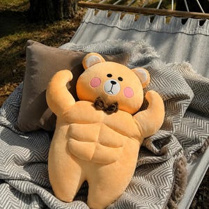 May include: A plush toy shaped like a light orange bear with a muscular torso featuring defined abdominal muscles. The bear has a small brown bow tie, pink circular cheeks, a white muzzle, and black embroidered eyes, nose, and mouth details.