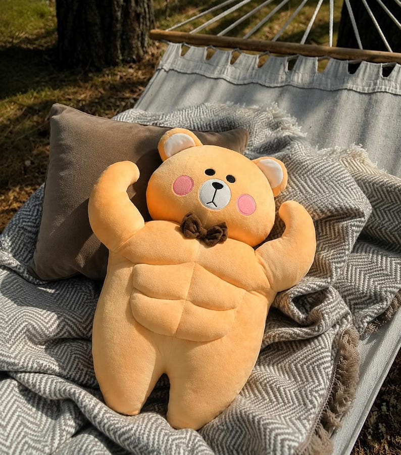 May include: A plush toy shaped like a light orange bear with a muscular torso featuring defined abdominal muscles. The bear has a small brown bow tie, pink circular cheeks, a white muzzle, and black embroidered eyes, nose, and mouth details.