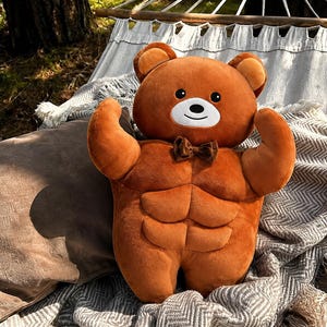 May include: A plush brown teddy bear toy featuring a muscular torso with defined abdominal muscles, rounded arms in a flexed pose, a white muzzle, black eyes, and a small brown bow tie attached at the neck. The bear has a soft, velvety texture.