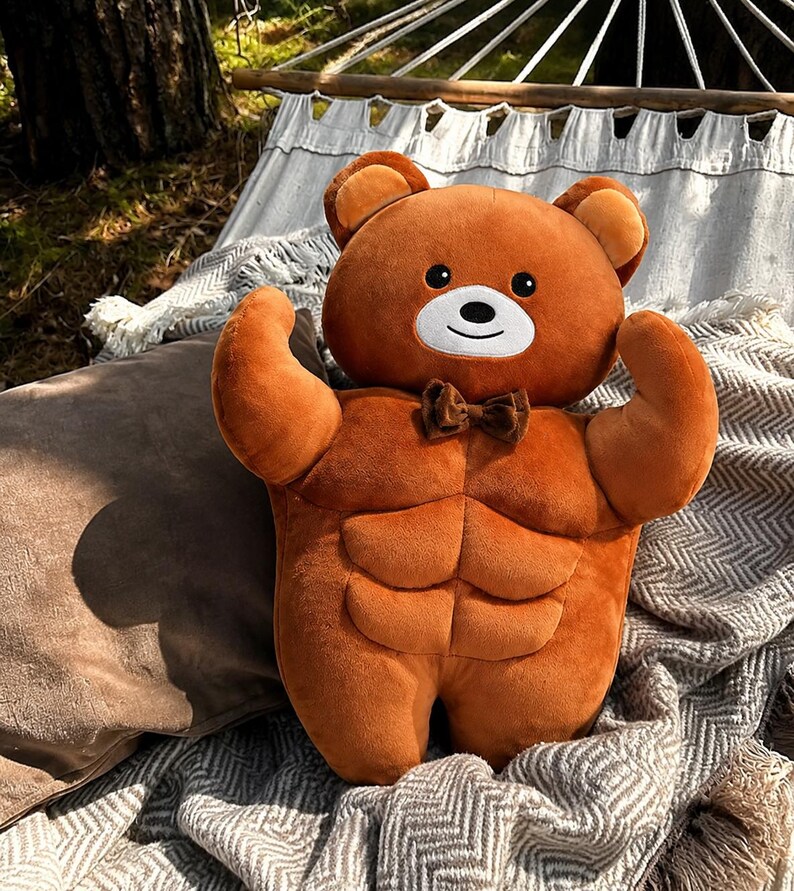 May include: A plush brown teddy bear toy featuring a muscular torso with defined abdominal muscles, rounded arms in a flexed pose, a white muzzle, black eyes, and a small brown bow tie attached at the neck. The bear has a soft, velvety texture.