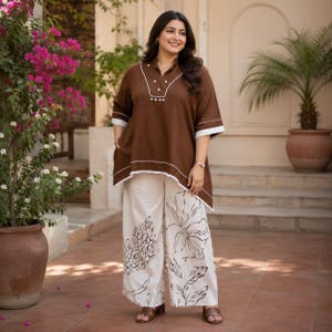 May include: A brown tunic with white trim and a V-neckline featuring small white buttons, paired with wide-leg white trousers decorated with a large black floral print. The tunic has three-quarter length sleeves with white cuffs and a hemline.